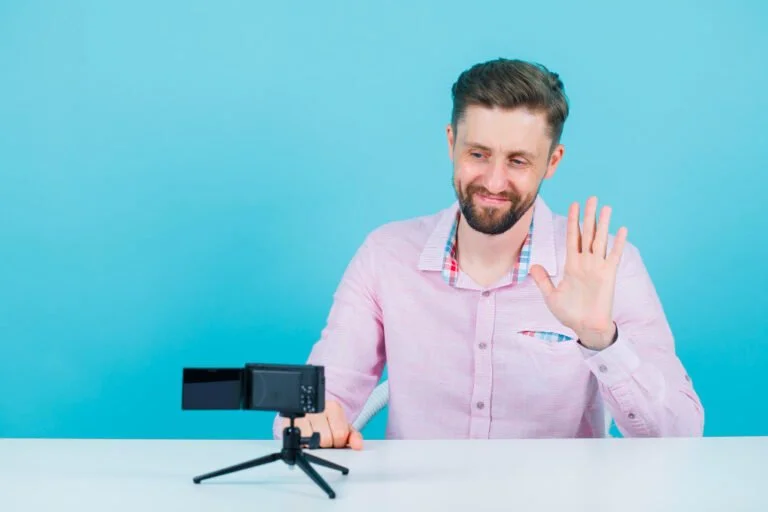 Dicas para criar vídeos profissionais com baixo orçamento 4 smiling blogger man is showing hi gesture by sitting front his mini camera blue background